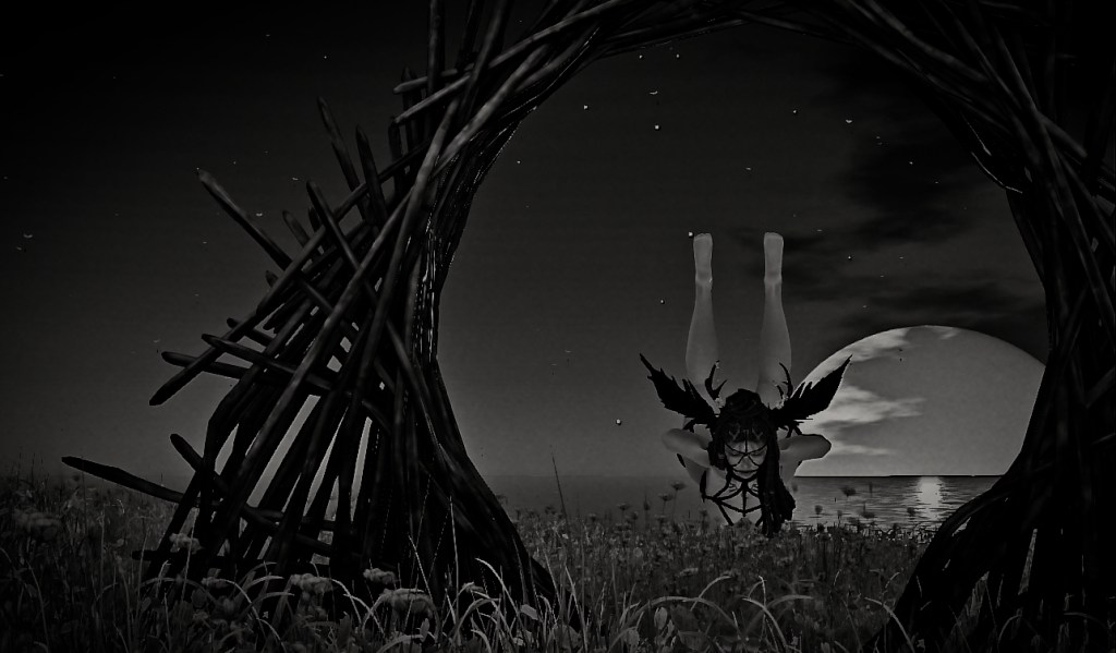 Black-and-white image of an avatar suspended upside down in a ritual pose, framed by a woven wooden arch under a night sky. A large moon rises over the ocean in the background, casting light across a field of flowers.
